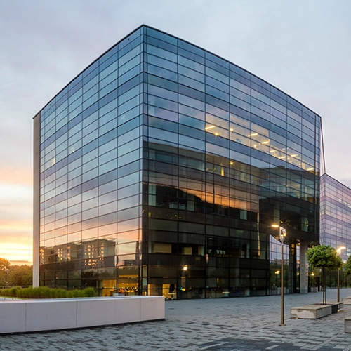 Modern glass office building with reflective windows at sunset, featuring lit interior lights, nearby trees, and a paved walkway with benches and street lamps in the foreground.