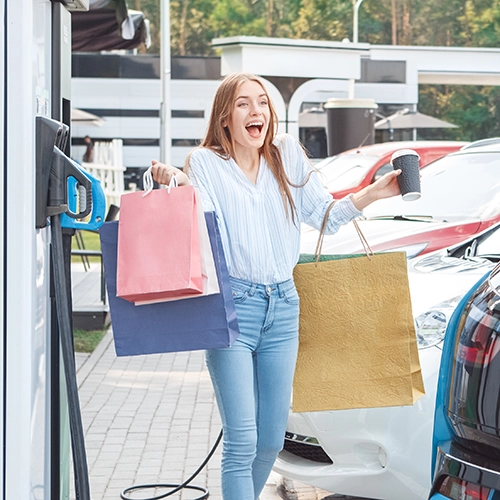 A smiling woman stands next to cars at a charging station, holding colorful shopping bags in one hand and a coffee cup in the other, appearing cheerful and excited.