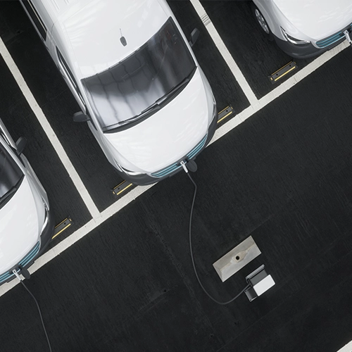 Overhead view of white electric cars parked in marked spaces, with one car plugged into a charging station on a dark asphalt surface.