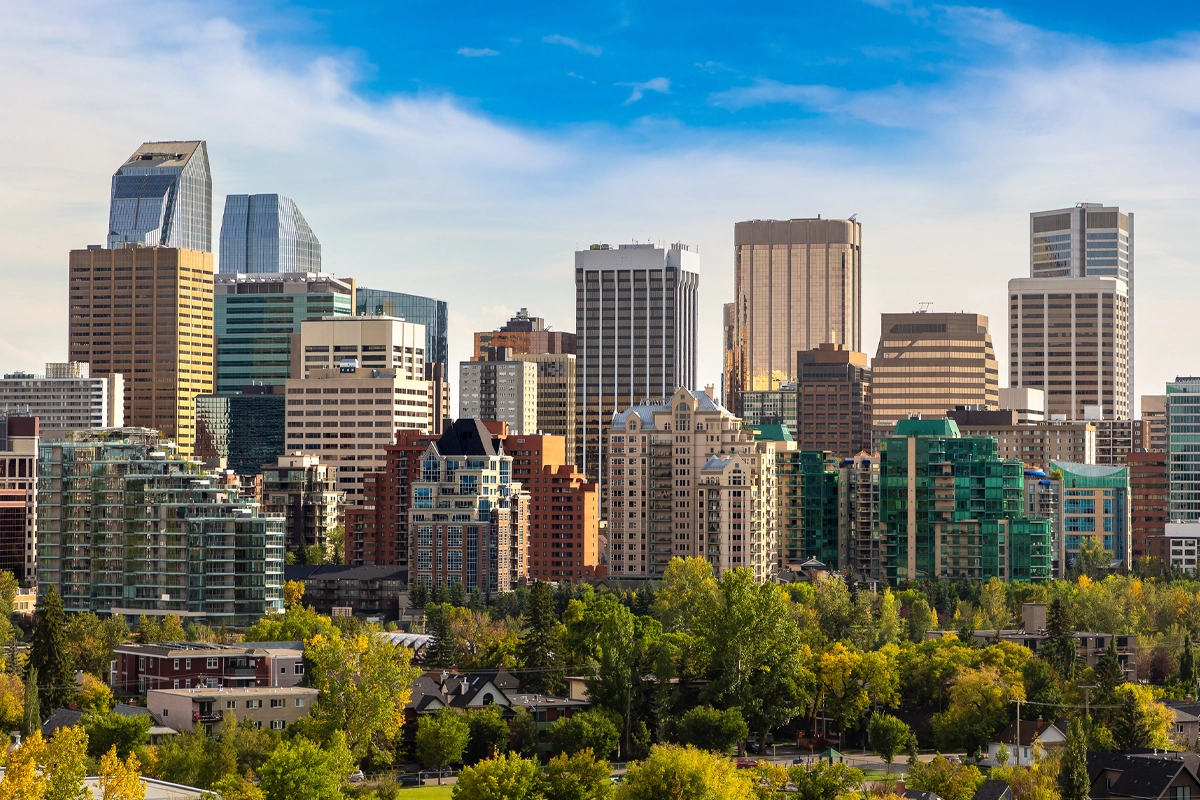 Downtown city skyline with modern high-rise buildings, glass towers, and green trees in the foreground under a bright blue sky—an ideal setting for Alberta EV charging funding to enhance sustainable urban living.