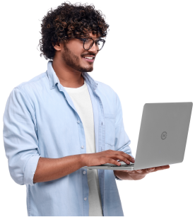 A young man with curly hair and glasses, wearing a light blue shirt over a white t-shirt, smiles while using a silver laptop, standing against a white background.